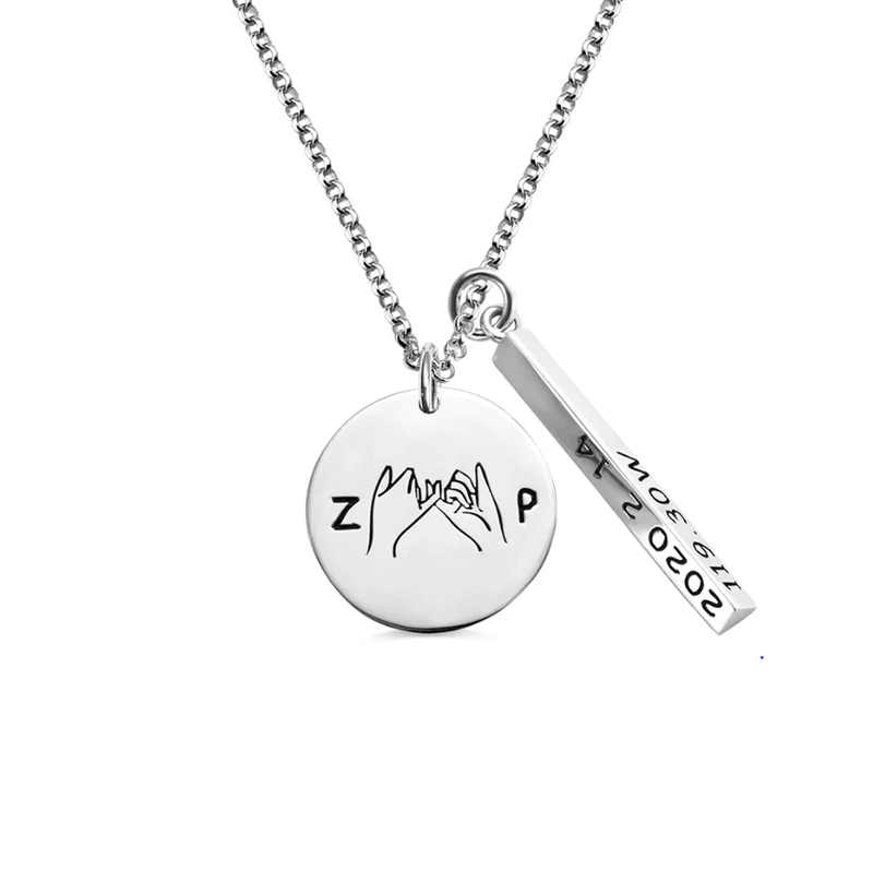 Silver necklace with two charms: one round charm featuring hand gestures forming a heart with "Z" and "P" letters, and a rectangular charm with engraved coordinates.