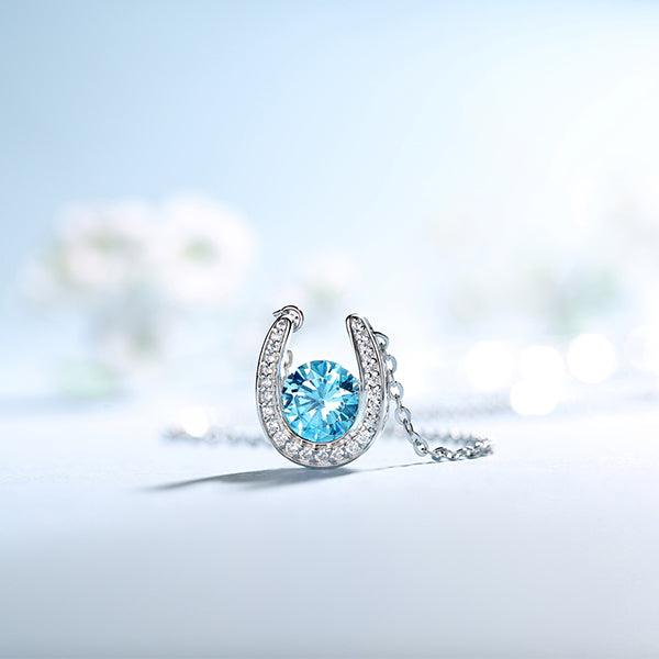 Sparkling silver horseshoe necklace with a blue gem, displayed against a soft-focus floral backdrop.