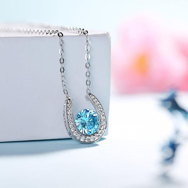 Chic horseshoe pendant with blue crystal on a chain, elegantly draped over a white box with a blurred floral background.