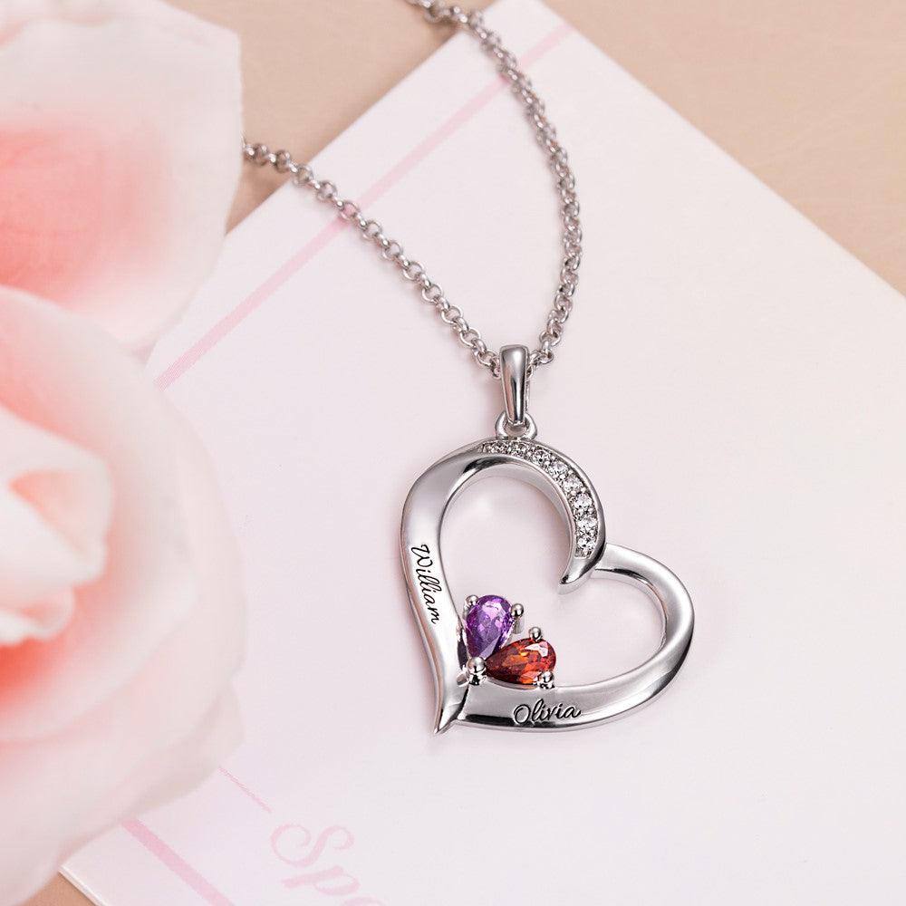 A heart-shaped pendant with purple and red gemstones, engraved with the names "William" and "Olivia," on a silver chain, placed against a light background with pink flowers.