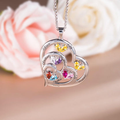 A heart-shaped pendant with colorful gemstones, each engraved with different names, on a silver chain, against a background of soft-focus roses.