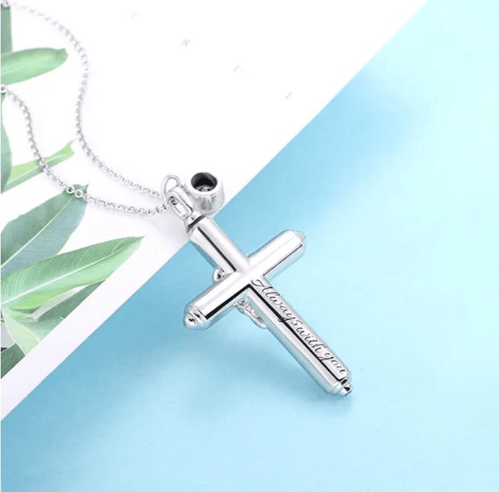 Silver cremation cross necklace with 'Always with you' inscription, displayed on a blue surface with green leaves