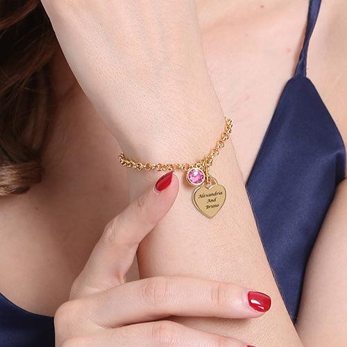 Woman wearing a gold-plated personalized heart charm bracelet with a pink birthstone and engraved names "Alexandria and Bruno," featuring an elegant link design.