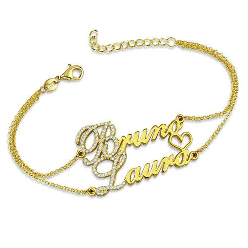 A gold bracelet with the names "Bruno" and "Laura" written in cursive, featuring birthstones just before the letters B and L, connected by a delicate chain and embellished with small diamonds and a heart.