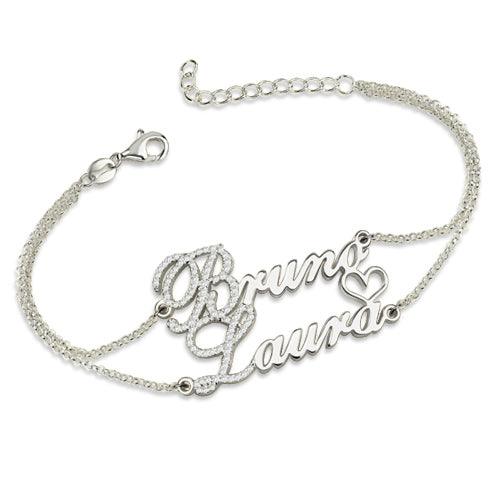 A  Silver bracelet with the names "Bruno" and "Laura" in cursive, featuring birthstones just before the letters B and L, connected by a delicate chain with small diamonds and a heart.