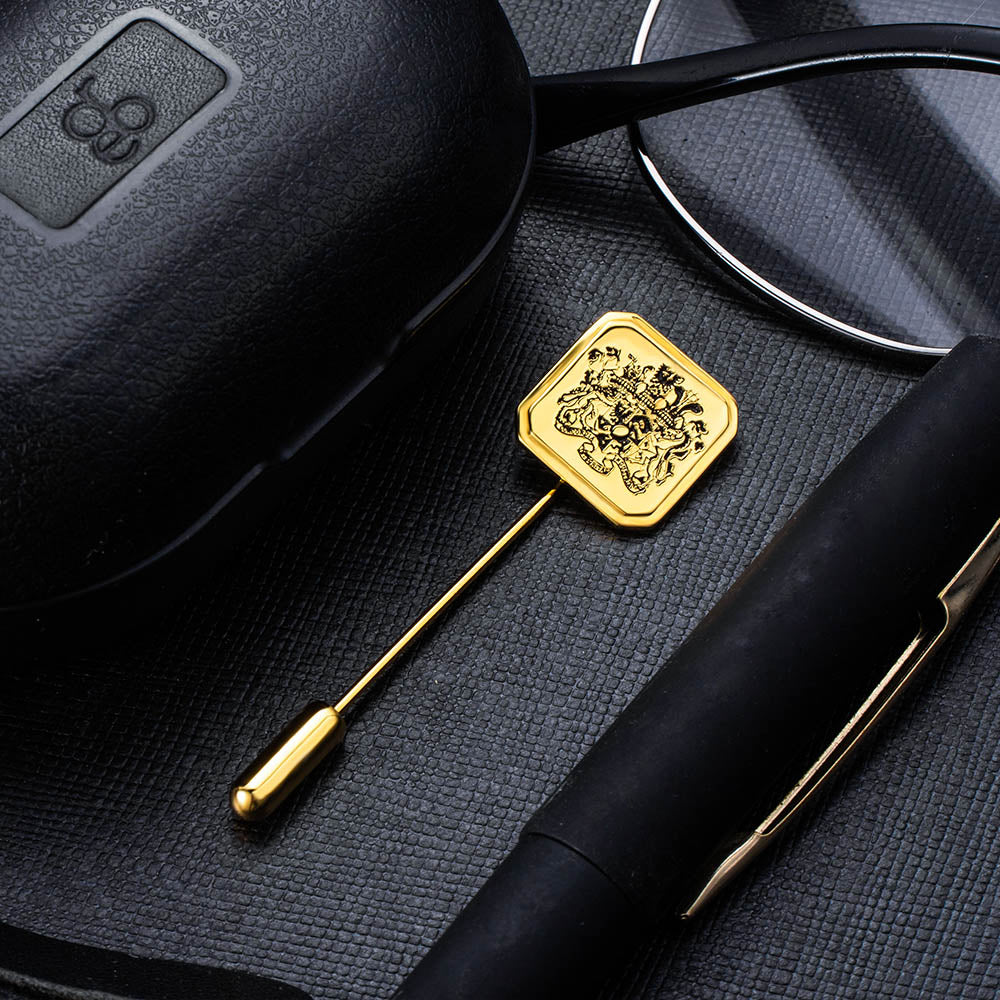 Gold hexagonal lapel pin with intricate family crest design, beside a pen and sunglasses on textured surface.