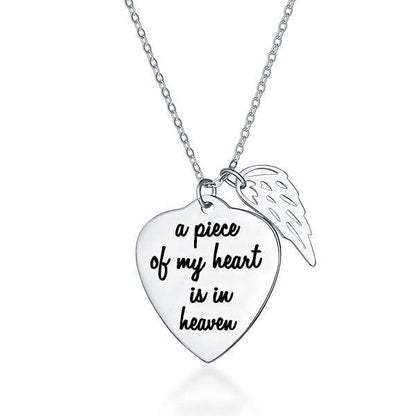 A silver necklace with a heart-shaped pendant engraved with "a piece of my heart is in heaven" and an angel wing charm attached.