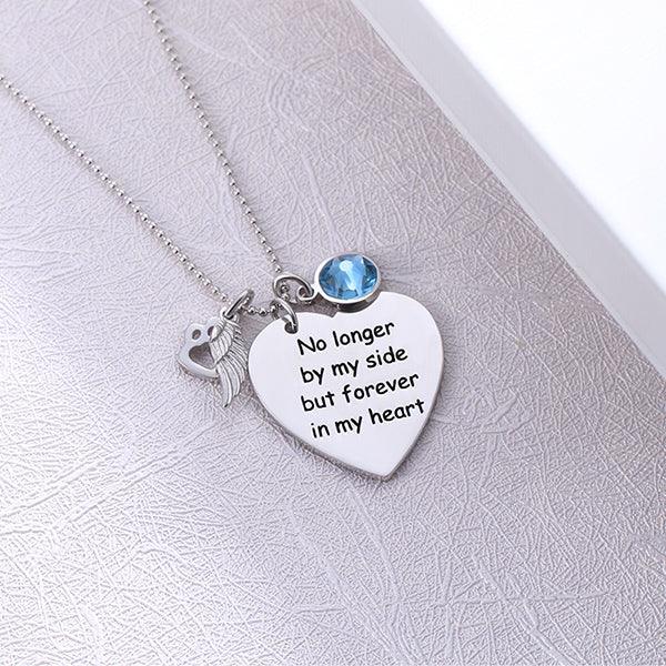 A silver necklace with a heart-shaped pendant engraved with "No longer by my side but forever in my heart," featuring a paw print, angel wing, and blue gem, laid on a textured surface.