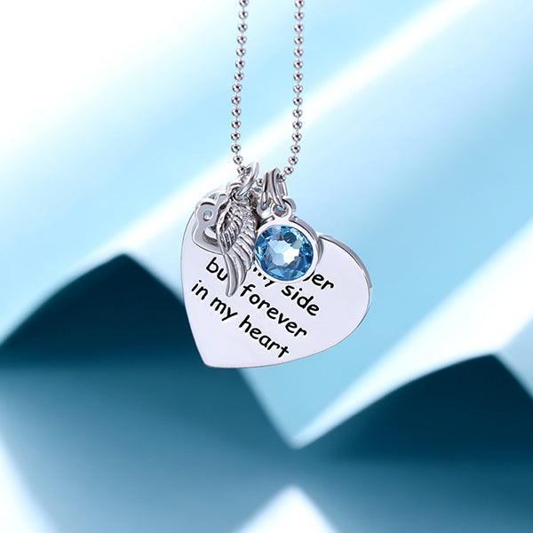 A silver necklace with a heart-shaped pendant engraved with "No longer by my side but forever in my heart," adorned with a paw print, angel wing, and blue gem, displayed against a light blue background.