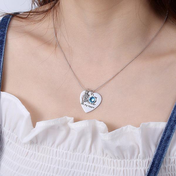 A woman wearing a silver necklace with a heart-shaped pendant engraved with "No longer by my side but forever in my heart," featuring a paw print, angel wing, and blue gem.