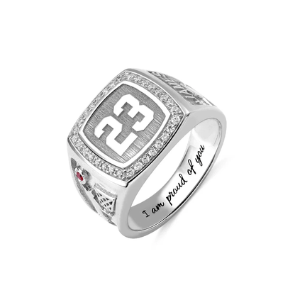 A silver championship ring with the number 23 prominently displayed, surrounded by small diamonds. The side features a red gem and a design, with "I am proud of you" engraved inside.