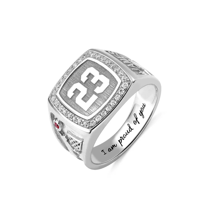A silver championship ring with the number 23 prominently displayed, surrounded by small diamonds. The side features a red gem and a design, with "I am proud of you" engraved inside.