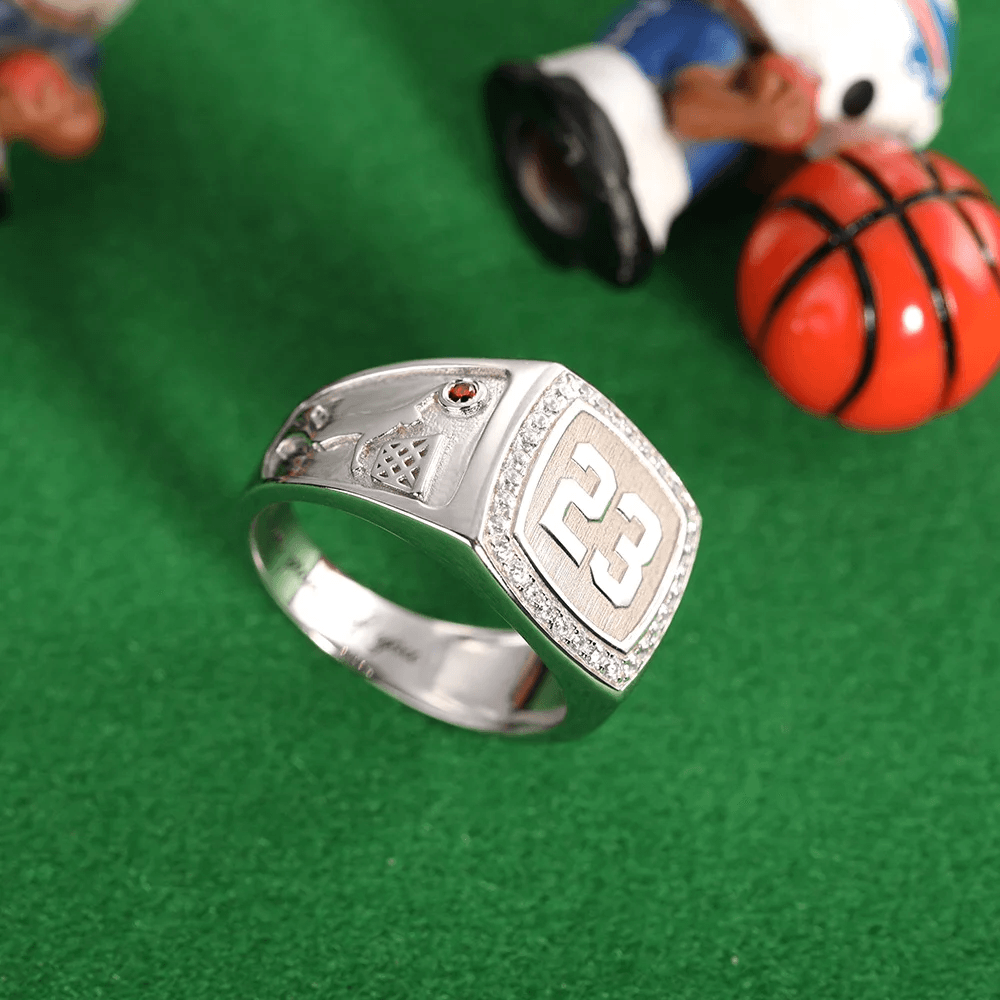 A silver championship ring featuring the number 23 surrounded by diamonds, with a red gem and basketball design on the side, set against a green background.