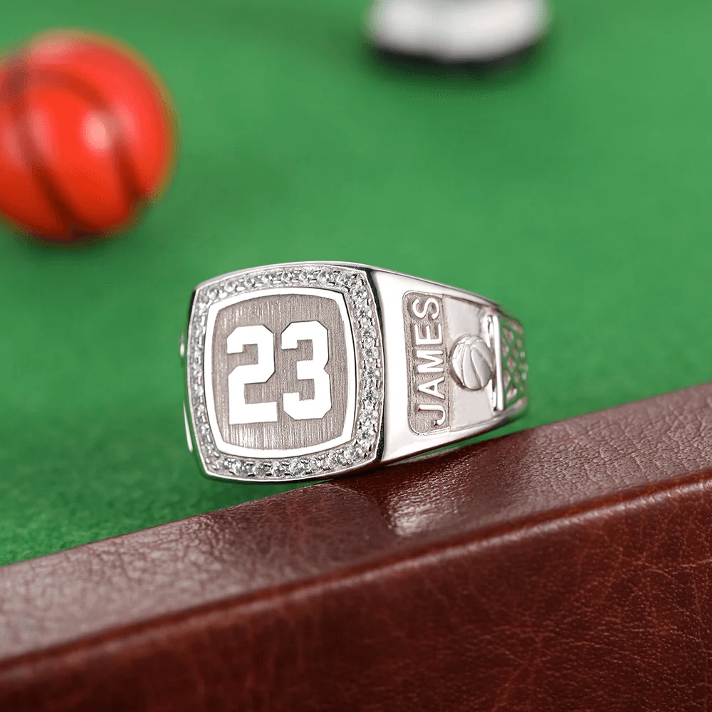 A silver championship ring with the number 23 surrounded by diamonds. The side features the name "JAMES" and a basketball design, set against a green background.