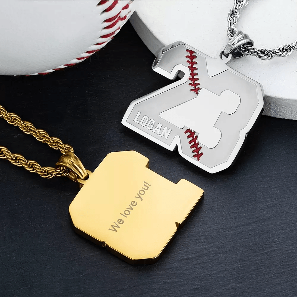 Two baseball-themed number pendants, one silver with "23" and "Logan" engraved, and one gold with "We love you!" engraved on the back, placed near a baseball.