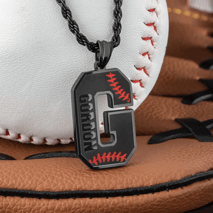 A black pendant shaped like the letter "G" with red baseball stitching details and the name "Gordon" engraved on it, placed on a black rope chain in front of a baseball glove and ball.