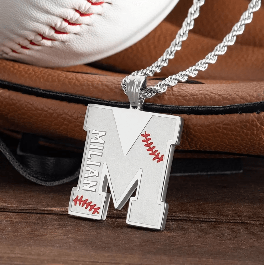 A silver pendant shaped like the letter "M" with red baseball stitching details and the name "Milian" engraved on it, placed on a silver rope chain in front of a baseball glove.
