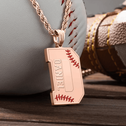 A rose gold pendant shaped like the letter "D" with red baseball stitching details and the name "Daniel" engraved on it, placed on a rose gold rope chain in front of a baseball glove and ball.