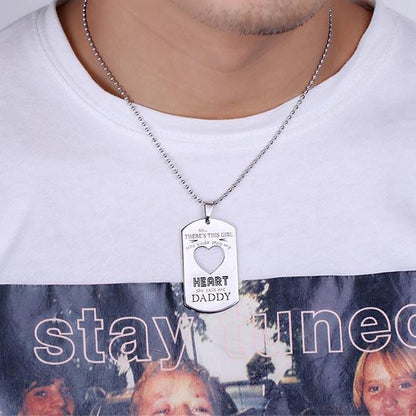 A man wearing a white shirt with a silver dog tag necklace that reads "she calls me DADDY" around a heart shape.
