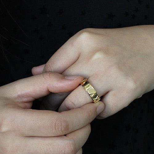 A person wearing a gold ring with engraved geometric patterns, seen against a dark background with a star pattern.