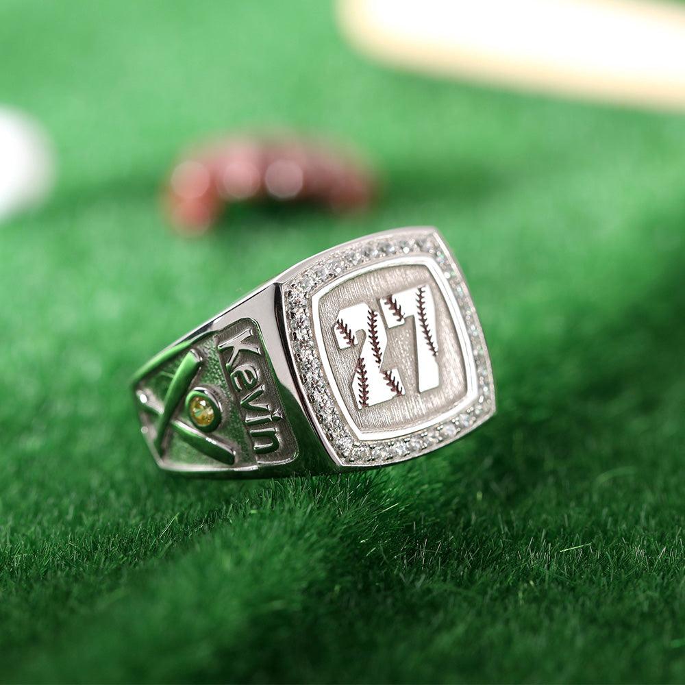 Customized silver baseball ring with name 'Kevin', green gem, and diamond-like border on a grassy background.