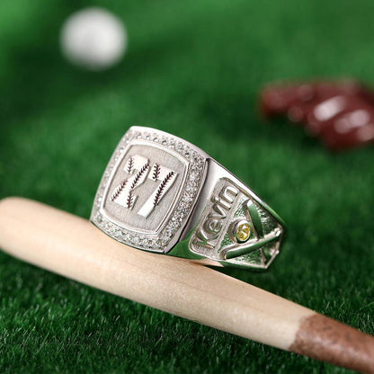 Engraved silver baseball ring with 'Kevin', stitching detail, and gem, on bat and grass backdrop.