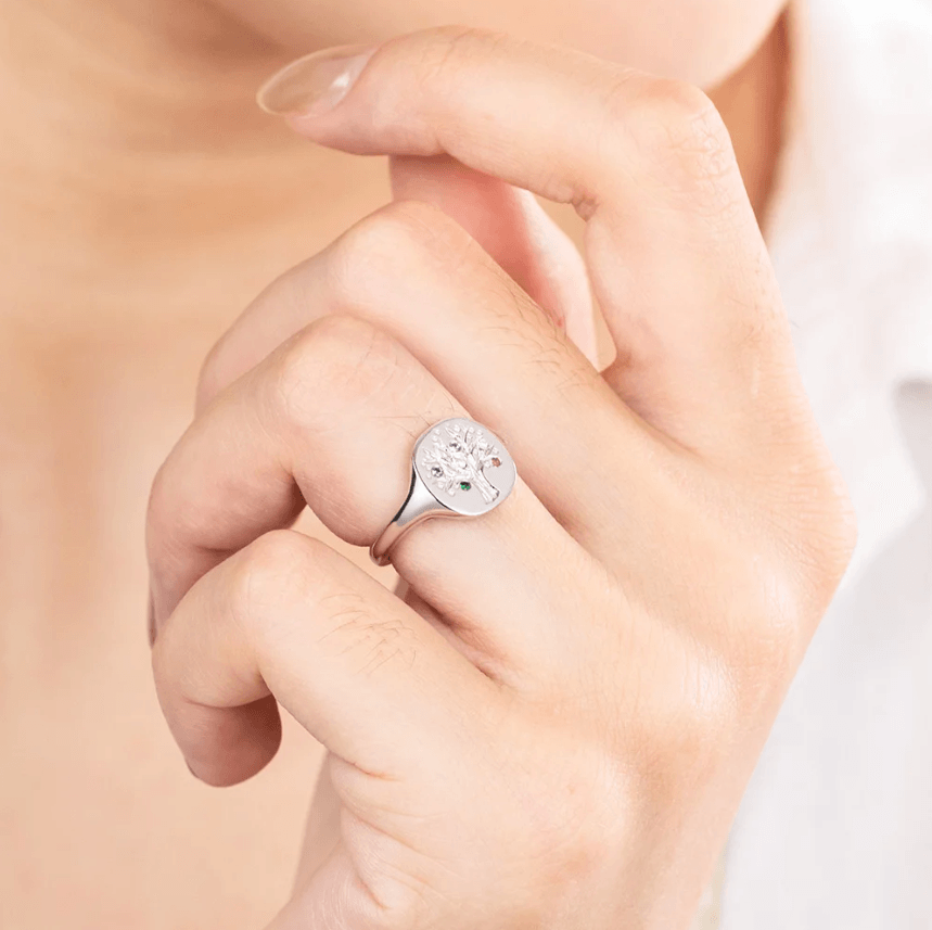 Fingers clutching a silver ring with a colorful tree design on a light background.