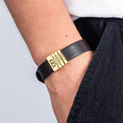 Man wearing a black leather bracelet with gold beads engraved 'Kaela', 'Celine', 'Anthony', on his wrist.