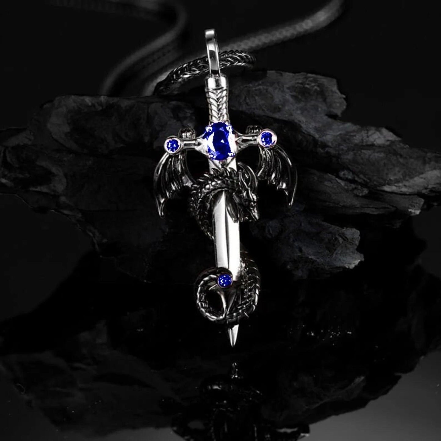 A pendant featuring a dragon entwined around a sword with blue gemstones, displayed on a foxtail chain against a dark background.