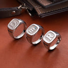 Three silver rings with engraved initials: "D" with "DAVID," "m" with a blue gem and "MY HERO," and "R" with "FRIEND," placed on a brown leather surface next to a wallet.