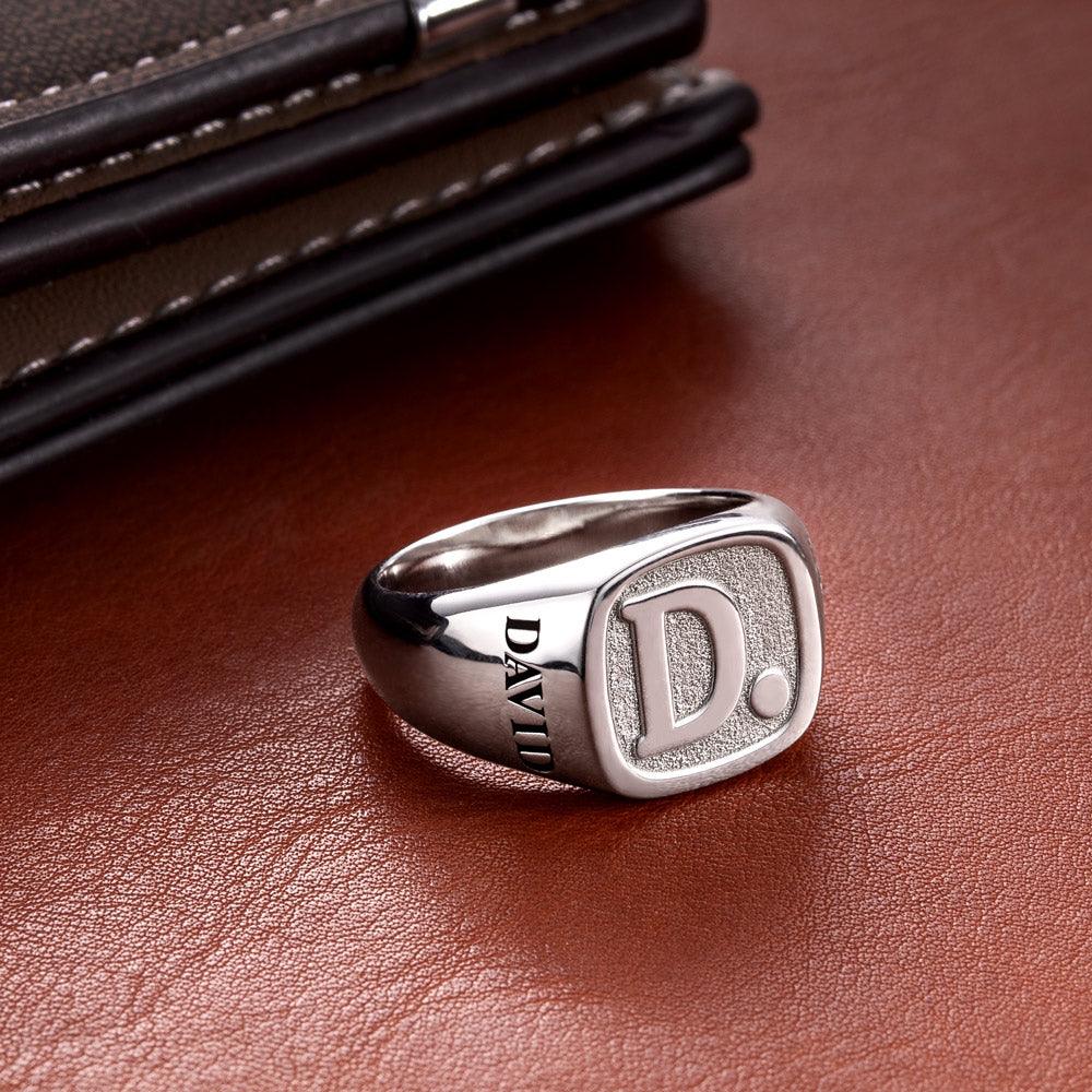 A silver ring engraved with the initial "D" and the name "DAVID," placed on a brown leather surface next to a black and brown leather wallet.