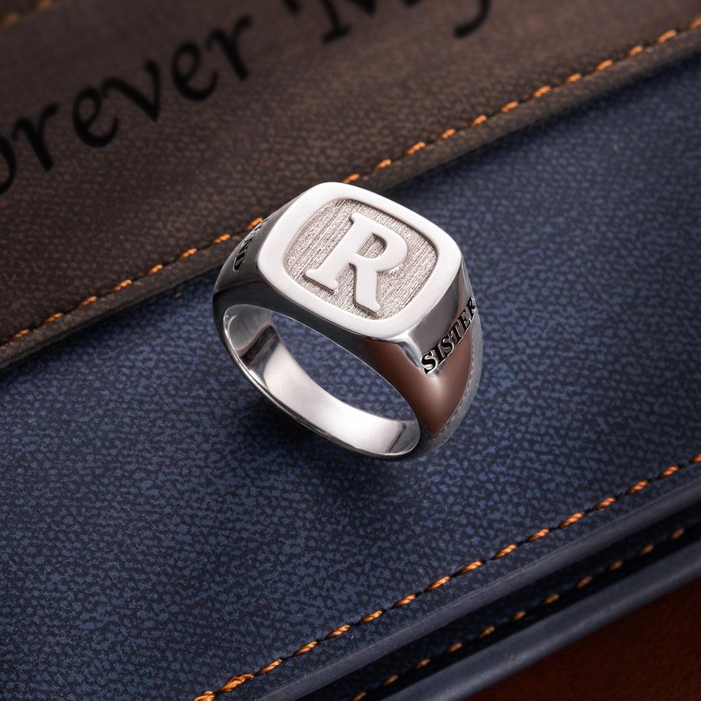 A silver ring engraved with the initial "R" and the word "SISTER," placed on a blue textured surface next to a brown leather item with orange stitching.