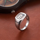 A silver ring engraved with the initial "m" and a blue gem, along with "MY HERO," placed on a brown leather surface next to a wooden watch.