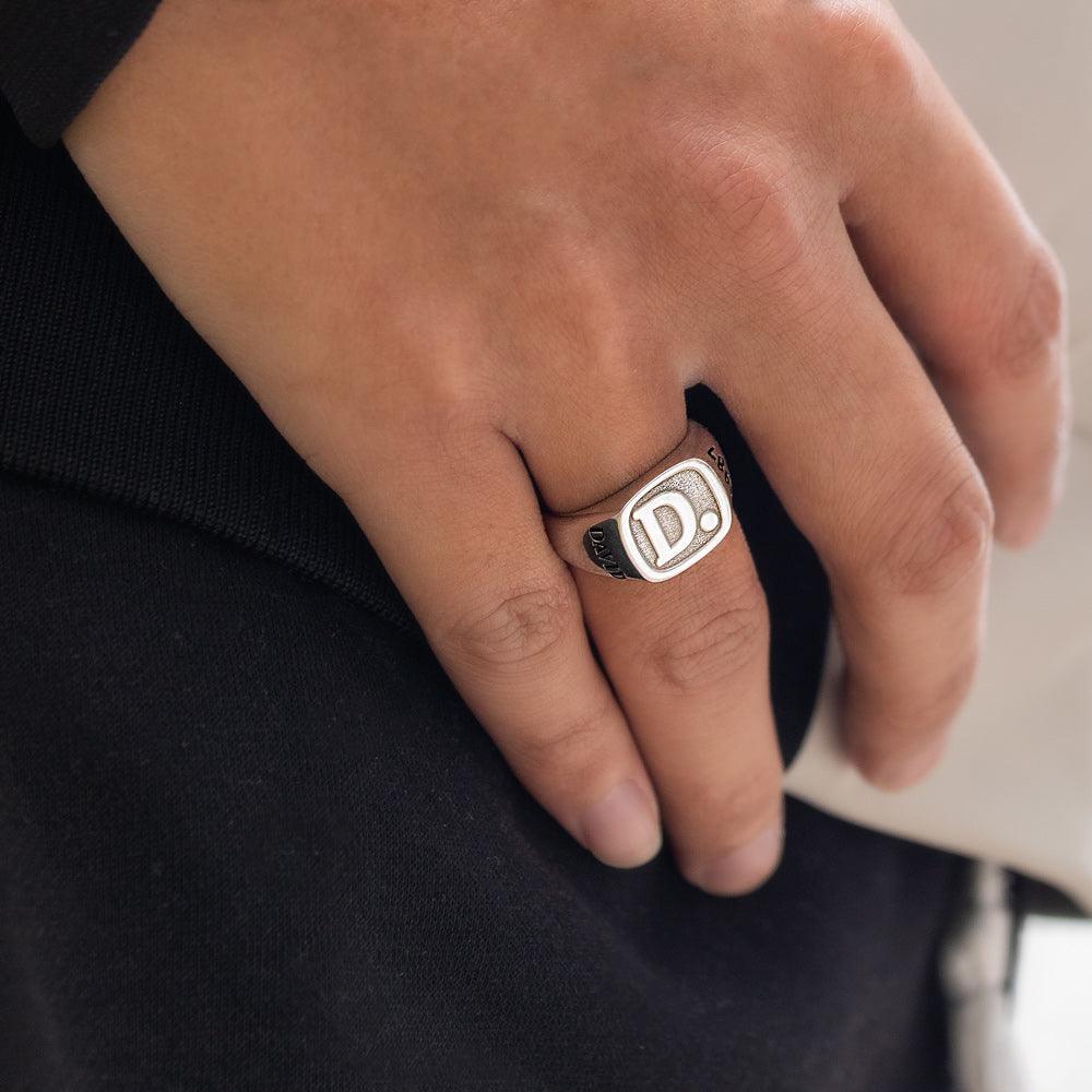 A person wearing a silver ring engraved with the initial "D" and the name "DAVID," shown on their finger, with a black and white outfit in the background.