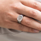 A person wearing a silver ring engraved with the initial "m," a blue gem, and the phrase "MY HERO," shown on their finger against a light-colored background.