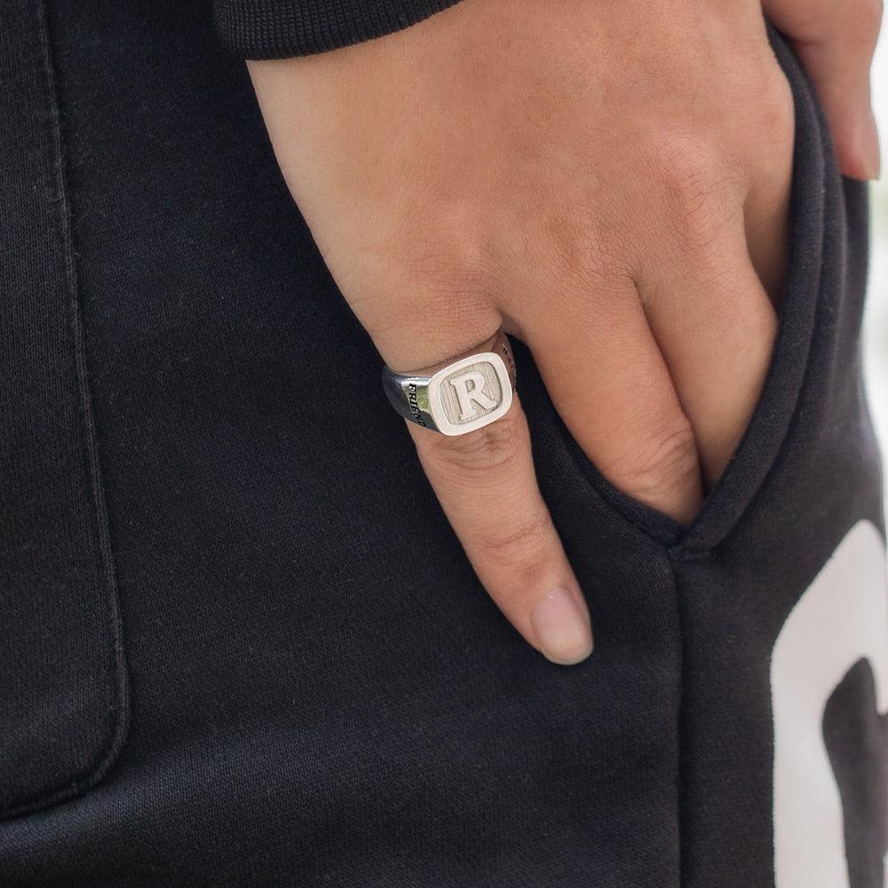 A person wearing a silver ring engraved with the initial "R" and the word "FRIEND," shown on their finger with their hand in the pocket of black pants.