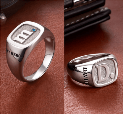 Two silver rings on a brown leather surface: one with the initial "m," a blue gem, and "MY HERO," and the other with the initial "D" and the name "DAVID."