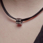 Close-up of a leather necklace with a cylindrical, silver pendant engraved with Roman numerals. The necklace rests against a person's neck.