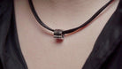 Close-up of a black leather necklace with a silver cylindrical pendant engraved with Roman numerals, worn around a person's neck.