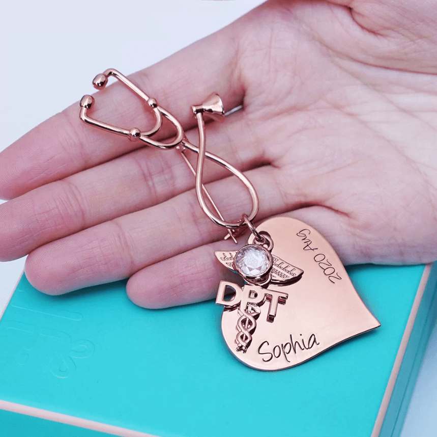 A close-up of a hand holding a rose gold nursing brooch with a stethoscope shape, medical emblem, and text reading "Sophia" and "Grad 2022."