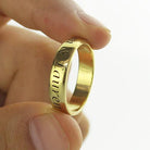 A hand holding a gold ring engraved with names "Lauren" and another partially visible name. The ring has a shiny and smooth surface.