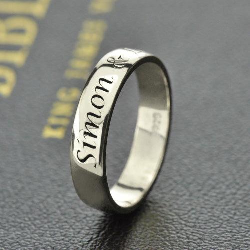 A silver ring engraved with the name "Simon" in an elegant script, displayed on a textured dark surface with faint gold text in the background.