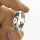 A hand holding a silver ring engraved with the names "Lauren" and another partially visible name. The ring has a shiny, smooth surface.