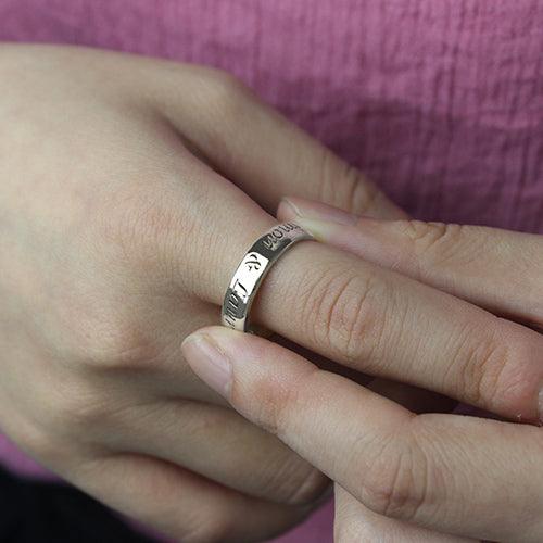 A person wearing a silver ring engraved with names, slipping it onto their finger. The backA person wearing a silver ring engraved with names, slipping it onto their finger. The background is a textured pink fabric.ground is a textured pink fabric.