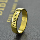 A gold ring engraved with the name "Simon" in an elegant script, displayed on a textured dark surface with faint gold text in the background.