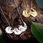 Silver and gold sloth-themed necklaces with personalized name engravings, displayed on a piece of wood and surrounded by leaves.