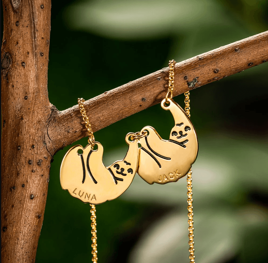 Gold sloth-themed necklace with two interconnected pendants, each engraved with a name, displayed on a wooden branch with a blurred green background.