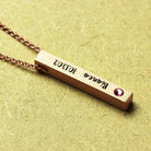 A gold necklace with a vertical bar pendant engraved with "Bianca 10.13.02" and a pink gemstone at the bottom left corner, displayed on a textured beige background.