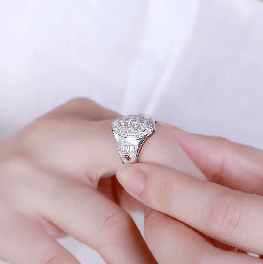 Person wearing a personalized 'KEVIN' football signet ring with a red birthstone.