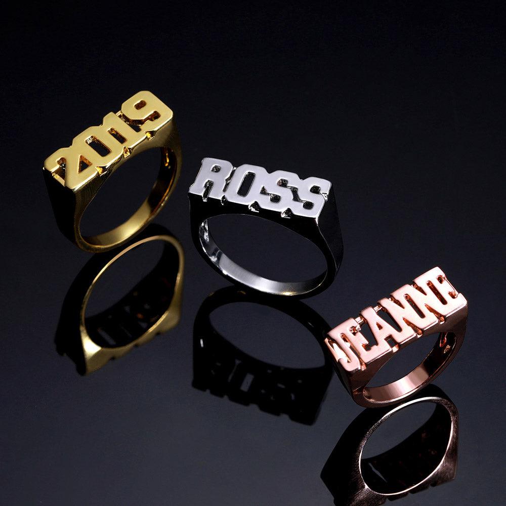 Gold '2019', silver 'ROSS', and rose gold 'JEANNE' block name rings on a dark background.